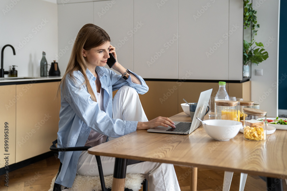 Busy woman freelancer has smartphone conversation working on laptop on home kitchen. Freelance, remote, distant work concept. Young caucasian female businesswoman doing job on computer calling phone. Busy woman freelancer has smartphone conversation working on laptop on home kitchen. Freelance, remote, distant work concept. Young caucasian female businesswoman doing job on computer calling phone.