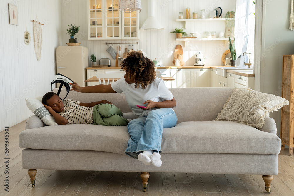 Tired small child fell asleep in VR glasses at home, African American mother taking off virtual reality helmet off sleeping nearby son kid. VR motion sickness in young children Tired small child fell asleep in VR glasses at home, African American mother taking off virtual reality helmet off sleeping nearby son kid. VR motion sickness in young children
