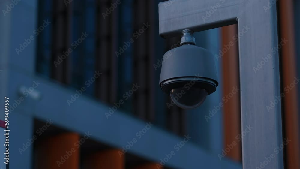 The silhouette of a round black CCTV camera, mounted on a pole against a dark corporate building The silhouette of a round black CCTV camera, mounted on a pole against a dark corporate building