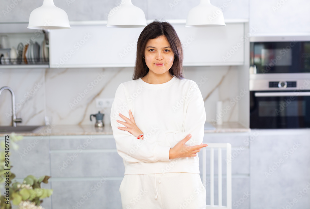 Smiling young brunette in white casual clothes standing in cozy light home kitchen interior..