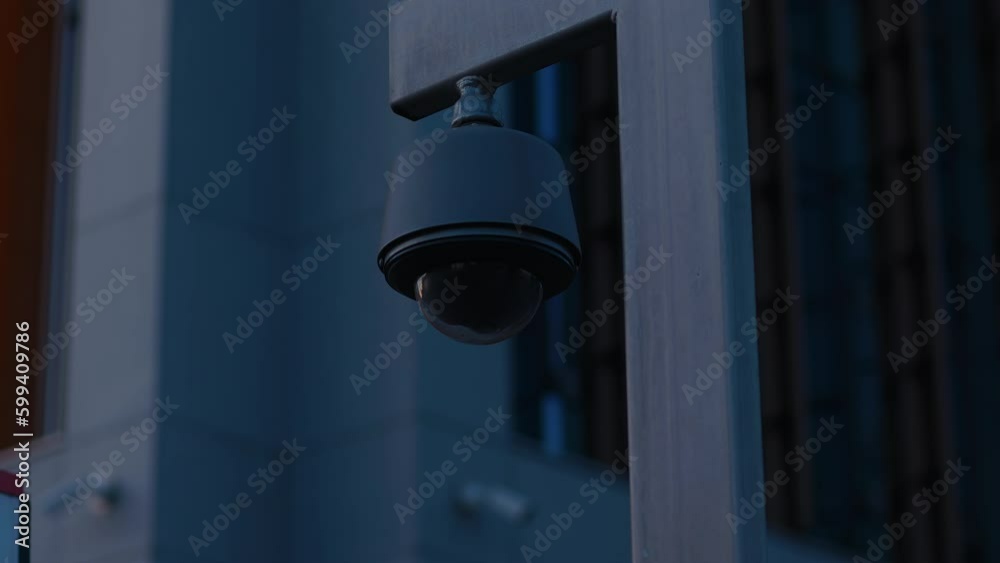 A pole-mounted round black CCTV camera silhouetted against a dark corporate building A pole-mounted round black CCTV camera silhouetted against a dark corporate building