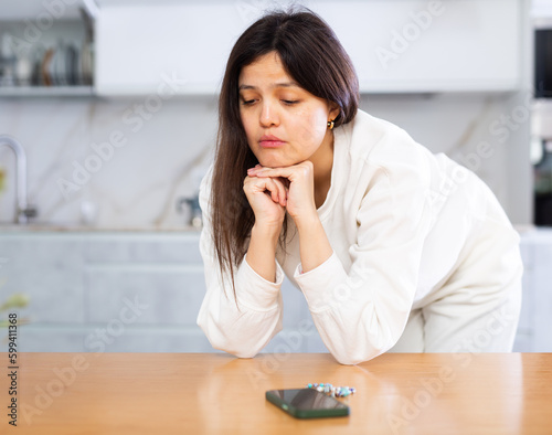 Sad upset young dark-haired woman waiting for phone call, standing leaning on table in home kitchen and staring at smartphone lying in front of her..