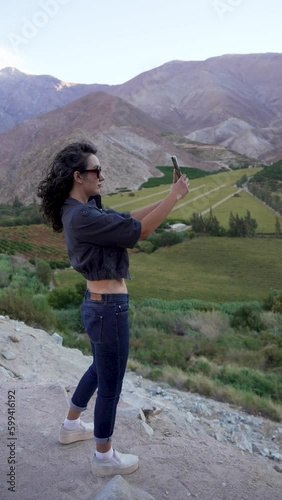 joven mujer latina de perfil grabando o tomando fotos con su celular al paisaje al atardecer en el Valle del Elqui Provincia de Paihuano photo