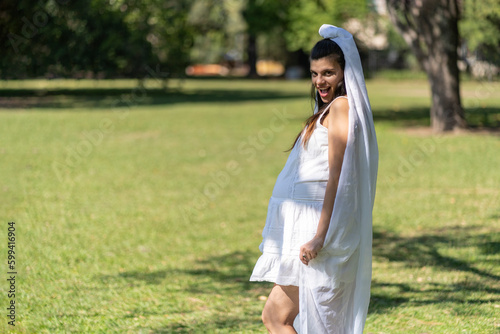 Pregnant bride in a white wedding dress holds her belly with her hand in a park. Seductive expression