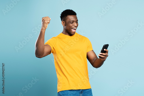 Happy African American man holding  mobile phone sports betting, win money isolated on blue background. Overjoyed gambler playing mobile game celebration success. Happy guy shopping online with sale