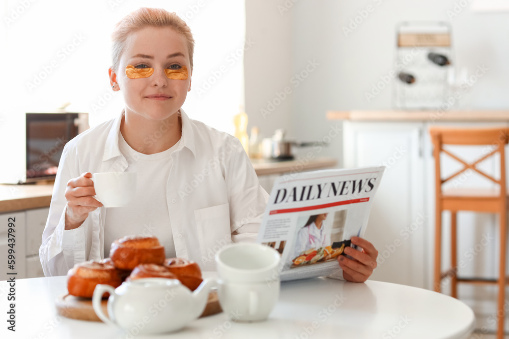 Young woman with cup of coffee reading newspaper in kitchen