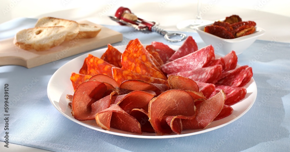 meat slices on a plate. traditional spanish sausage with beef jerky. salchichon, chorizo and prosciutto on a light background. serving cold cuts with dried tomatoes and bread. antipasto in sunlight. meat slices on a plate. traditional spanish sausage with beef jerky. salchichon, chorizo and prosciutto on a light background. serving cold cuts with dried tomatoes and bread. antipasto in sunlight.