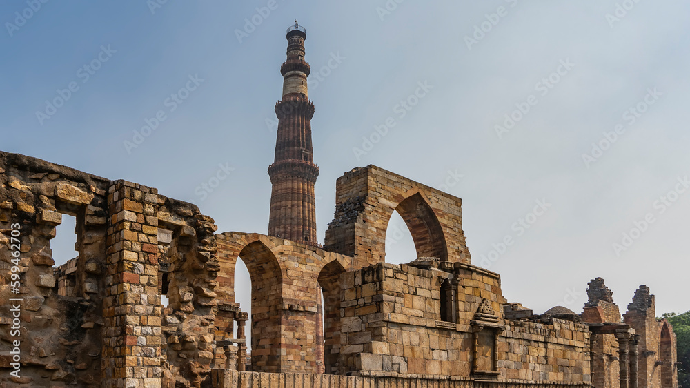 Ruins of the ancient temple complex Qutab Minar. Dilapidated walls with arches, carved decorations. Pigeons sit on ledges. The world's tallest brick minaret stands against the blue sky. India. Delhi
