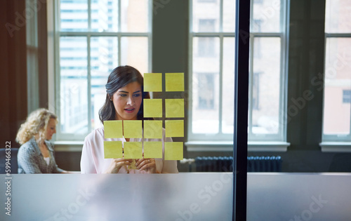 Turning a good idea into a great one. a businesswoman reading adhesive notes on a glass wall with a colleague in the background.