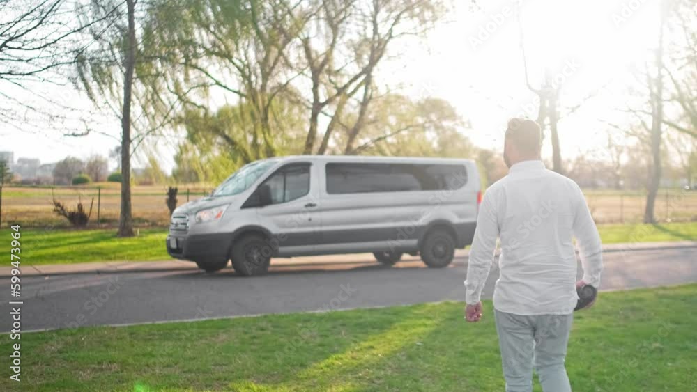 Adult man walking towards a large van during sunset wearing casual clothes with weeping willows in the background Adult man walking towards a large van during sunset wearing casual clothes with weeping willows in the background