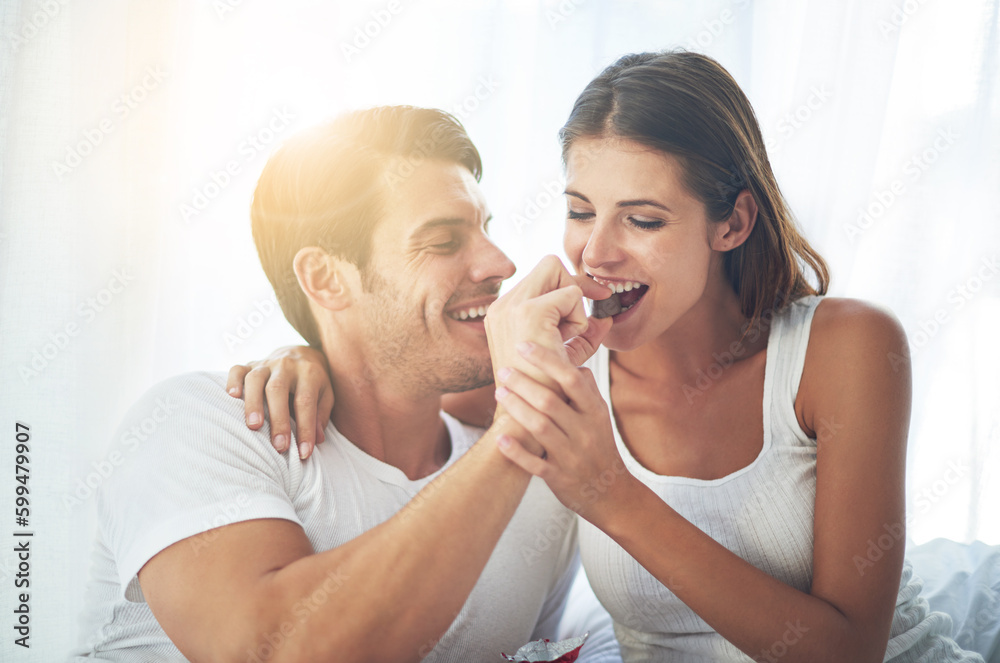 Love, happy couple eat chocolate in bedroom and care with smiling or laughing. Valentines day, married people sharing sweet snack and spending quality time in their home and romance or caring