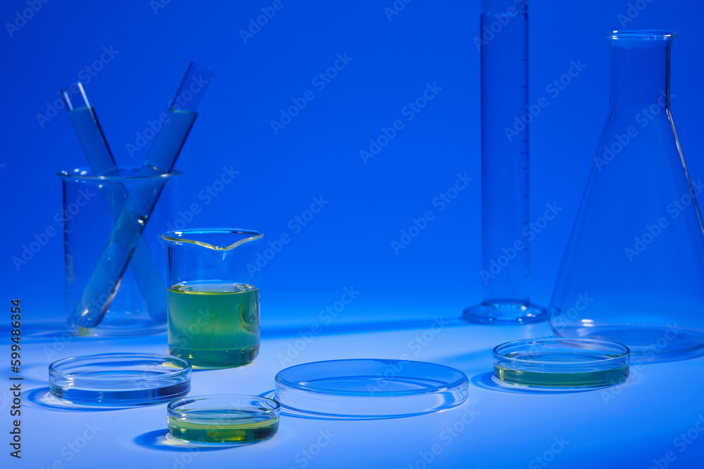 Beaker and petri dish of yellow liquid displayed with other laboratory glassware on a blue background. Vacant space on podium Beaker and petri dish of yellow liquid displayed with other laboratory glassware on a blue background. Vacant space on podium