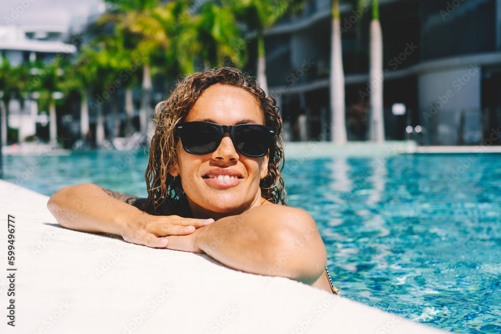 Happy woman enjoy sun and pool in hotel resort scenic holiday place. People on summer vacation. Travel and luxury lifestyle. Young lady smiling on poolside and wearing sunglasses alone in blue water