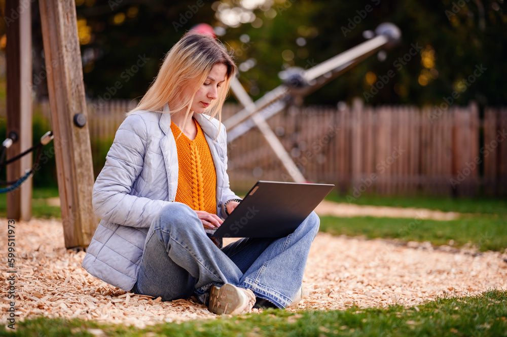 Young girl in spring outwear sitting on earth in playground near fence using laptop