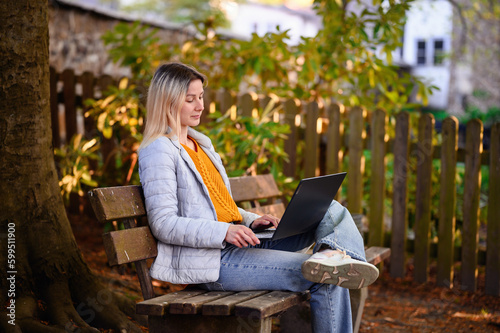 Young girl in spring outwear sitting on old wooden bench near tree and fence using laptop