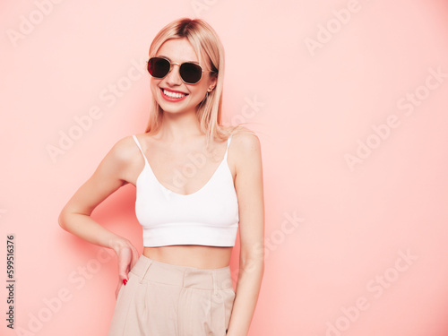 Young beautiful smiling blond female in trendy summer white top t-shirt and beige trousers clothes. Sexy carefree woman isolated on pink background in studio. Positive model having fun. Sunglasses © halayalex