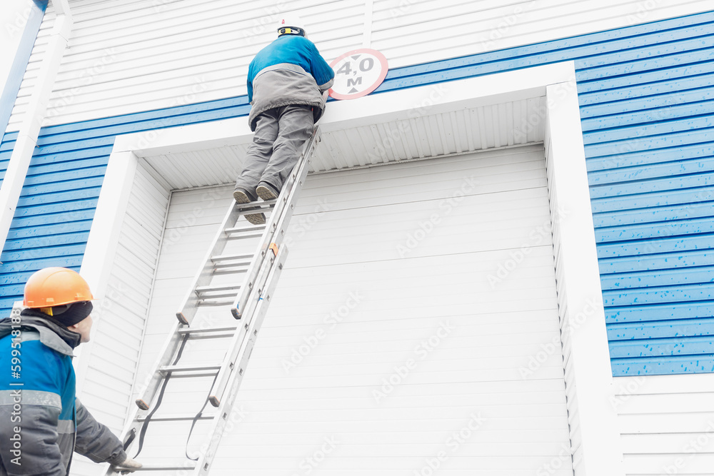 Two construction workers fix sign on automatic garage door for large vehicles. Worker stands on ladder and sets up height limit sign for entering garage. Two construction workers fix sign on automatic garage door for large vehicles. Worker stands on ladder and sets up height limit sign for entering garage.