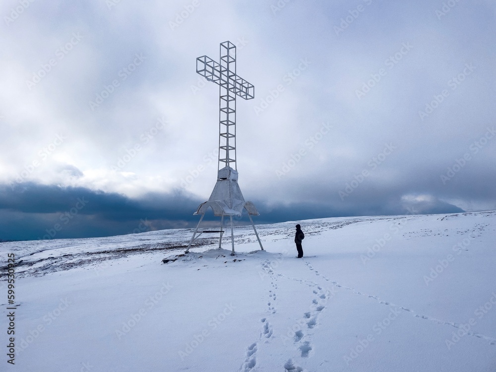 Man with backpack standing on a field covered in snow close to a tall steel cross Man with backpack standing on a field covered in snow close to a tall steel cross