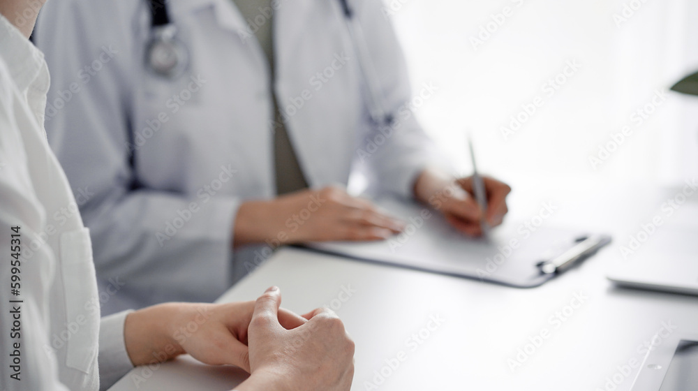 Doctor and patient sitting at the desk in clinic office. The focus is on female physician's hands using tablet computer, close up. Perfect medical service and medicine concept Doctor and patient sitting at the desk in clinic office. The focus is on female physician's hands using tablet computer, close up. Perfect medical service and medicine concept