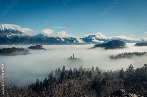 A mystical view from a viewpoint overlooking Bled lake, obscured by thick fog. The church and castle emerge like ethereal islands, surrounded by misty mountains in the background.