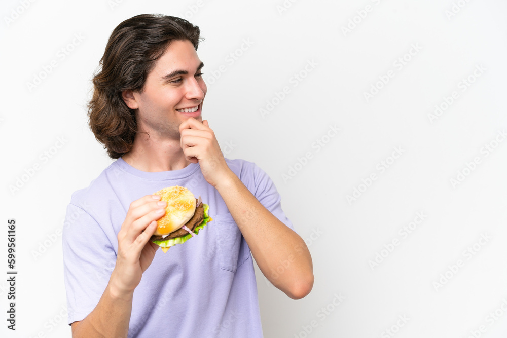 Young handsome man holding a burger isolated on white background thinking an idea and looking side Young handsome man holding a burger isolated on white background thinking an idea and looking side