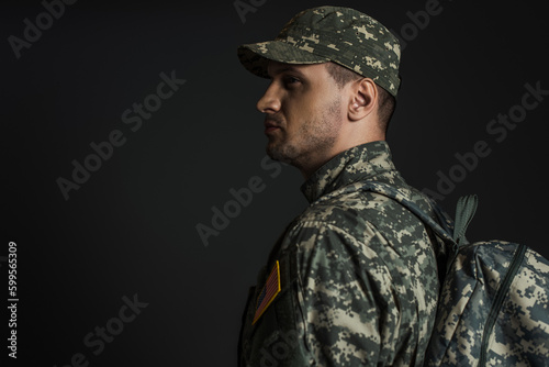 patriotic soldier in camouflage uniform standing with backpack isolated on black.