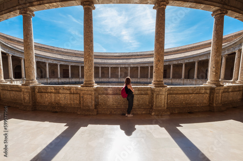 Young female tourist with backpack enjoying wide galleries with columns in the inner circular patio in Palace of Charles V in Alhambra complex in Granada, Andalusia, Spain.