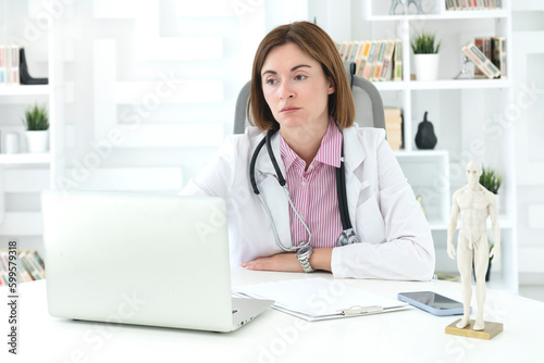 Young attractive female doctor sitting at the desktop in the office and consulting her patient online 