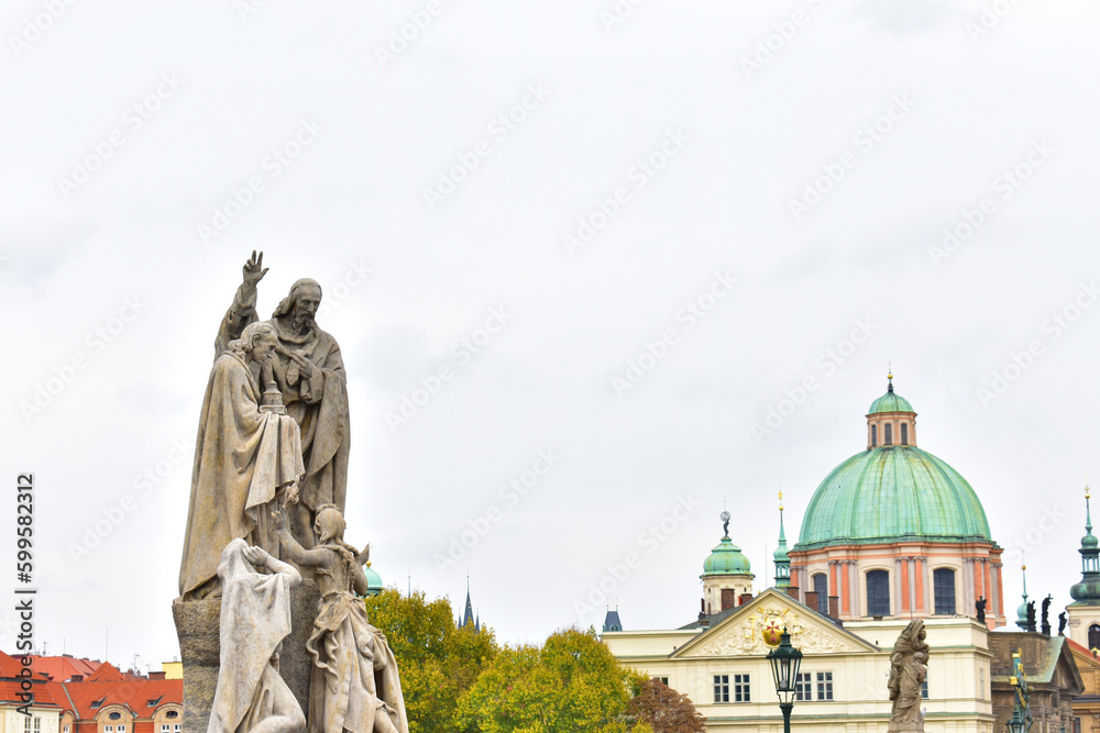 Ancient catholic sculptures on a pedestal and old buildings with green dome and towers with the green top. Statues on biblical subjects. Charles Bridge. Prague, Czech Republic, October 2022.