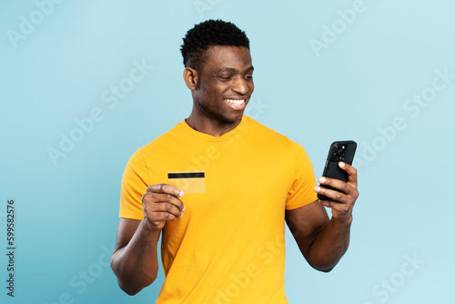 Portrait of handsome smiling African American man holding credit card, suing mobile app shopping online isolated on blue background. Happy stylish male ordering food. Technology concept 