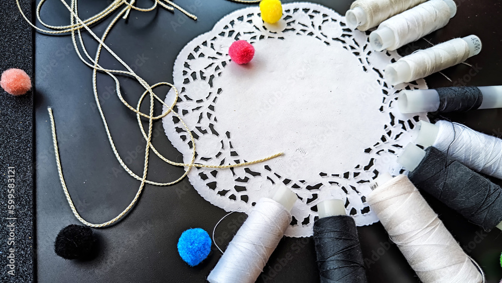 White and black spools of thread with decorative napkin and colored soft fluffy balls on a black background. Black card with abstract frame, texture and sewing threads. Top view. Partial focus White and black spools of thread with decorative napkin and colored soft fluffy balls on a black background. Black card with abstract frame, texture and sewing threads. Top view. Partial focus
