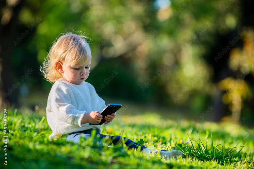 Kid holding a phone in his hands while outdoors. A Caucasian child staring into a smartphone with interest, sitting in a park alone on a sunny day. Cute child sitting in a park on a nice sunny day. Kid holding a phone in his hands while outdoors. A Caucasian child staring into a smartphone with interest, sitting in a park alone on a sunny day. Cute child sitting in a park on a nice sunny day.