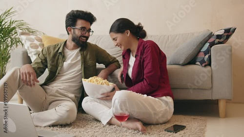 Happy cheerful indian couple watching laptop movie and eating popcorn on floor