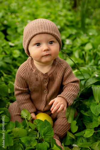 a surprised little boy (baby) in a brown suit and hat is sitting in the green grass and holding a green apple in his hands