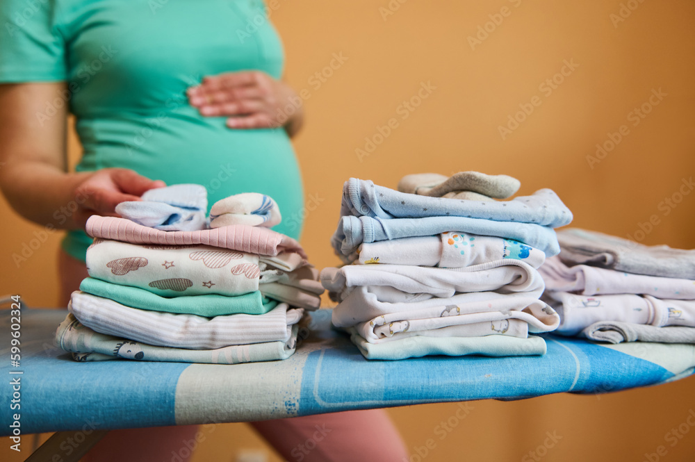 A stack of folded ironed clean baby clothes on ironing board, against the background of blurred pregnant woman in turquoise t-shirt, gently caressing her belly. Pregnancy. Preparation to childbirth A stack of folded ironed clean baby clothes on ironing board, against the background of blurred pregnant woman in turquoise t-shirt, gently caressing her belly. Pregnancy. Preparation to childbirth
