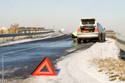 Road accident. Warning triangle and driver in a reflective safety vest changing the car wheel. Focus on triangle