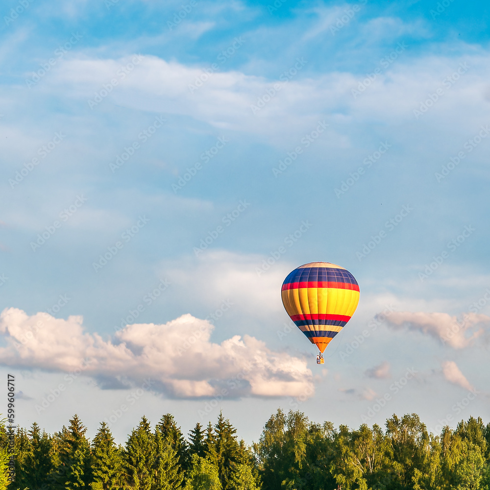 A bright hot air balloon flies in the blue sky above the treetops of the forest. Bright warm colors