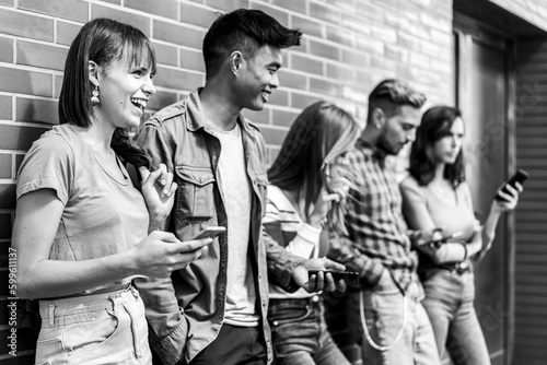 Multicultural friends laughing using smartphone at wall on university college campus - Young people addicted by mobile smartphones - Technology concept with always connected milenials - Vintage edit photo