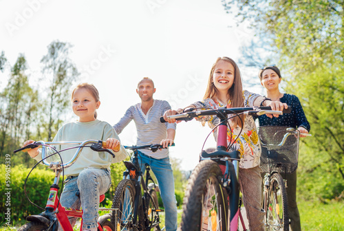 Smiling father and mother with two daughters during summer outdoor bicycle riding. They enjoy togetherness in the summer city park. Happy parenthood and childhood or active sport life concept image.