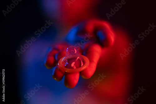 Male hand holding and offering condom. With red and blue colors predominant in the image. Nightclub or party. With blurred dark background.