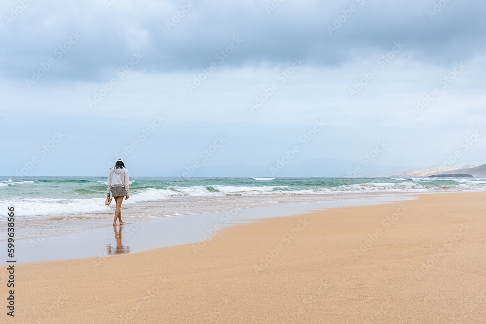 Mujer caminando en playa solitaria de cielo nublado con  chanclas en las manos y sudadera