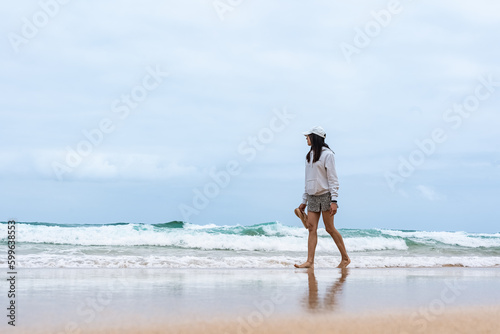 Mujer caminando en playa solitaria de cielo nublado con chanclas en las manos y sudadera