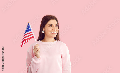Cute smiling female american citizen with usa national flag isolated on pink background. Happy young woman celebrates America's Independence Day and with flag in hand smiling looks away at copy space.