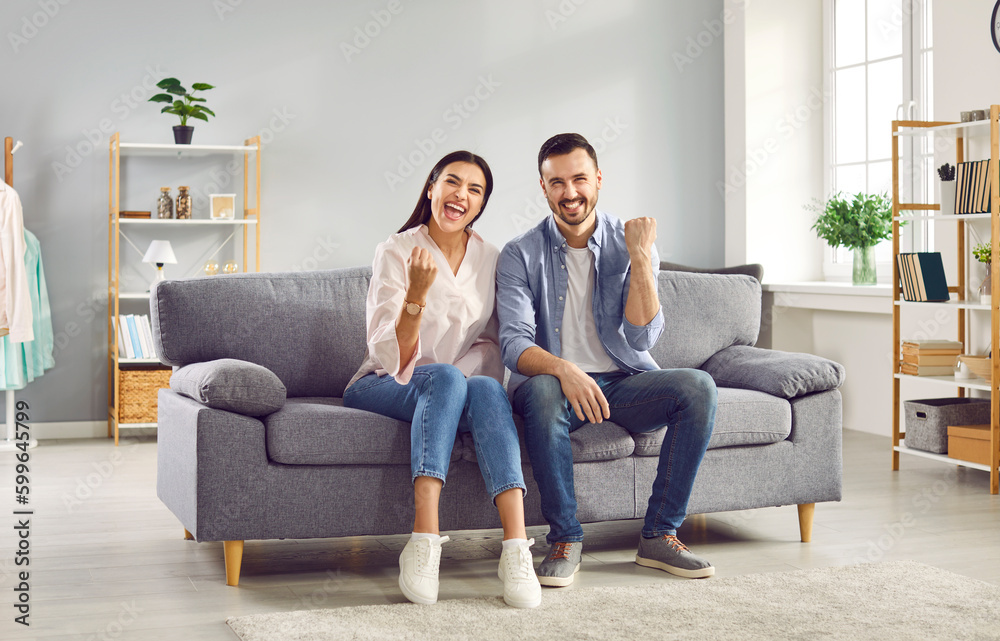 Excited couple raising fists in excitement while sitting on sofa at home. Overjoyed happy young man and woman watching soccer or hockey match on TV, celebrating victory of their team