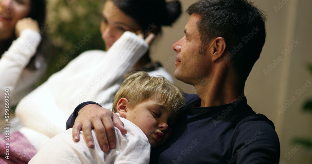 Young father holding sleeping child at night while in conversation with friends, candid casual man with son Young father holding sleeping child at night while in conversation with friends, candid casual man with son