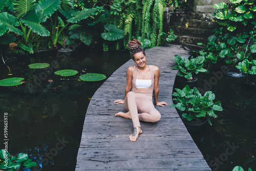 Portrait of cheerful Caucasian woman in trendy sportswear resting in tranquil nature environment for yoga meditation during summer retreat vacations in Indonesia  happy female posing at pond