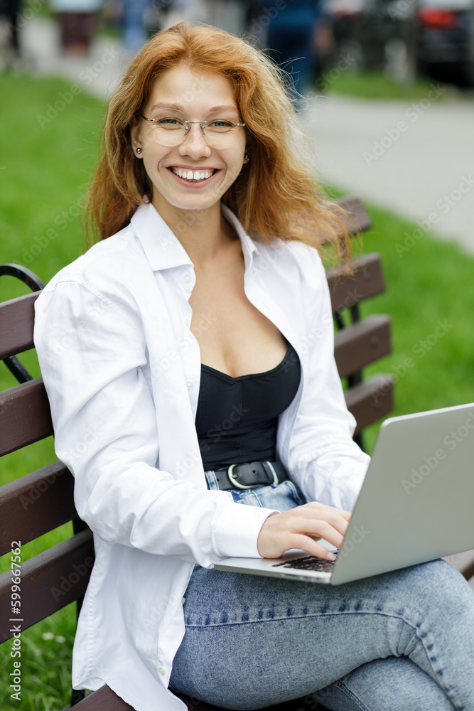 Young beautiful confident redhead girl student using her computer sitting on the bench in the park. Young beautiful confident redhead girl student using her computer sitting on the bench in the park.