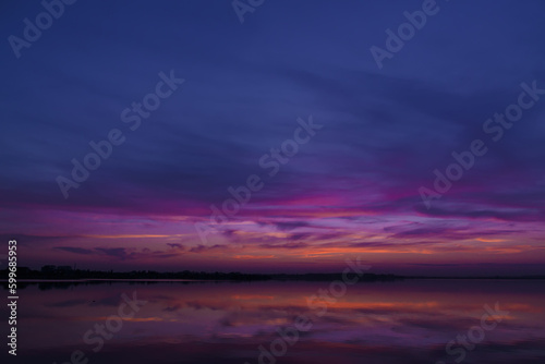 Fototapeta Naklejka Na Ścianę i Meble -  Scenic view of colorful sky reflected in the lake at evening, after sunset. Sunset at coast of the lake. Reflection, blue sky and yellow sunlight. Landscape during sunset. The beautiful nature around