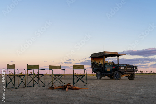 Ein grüner Safari Jeep steht bei Sonnenuntergang in einem ausgetrockneten Salzsee der Makgadikgadi Pan in Botswana, Afrika, daneben Campingstühle rund um ein Lagerfeuer photo