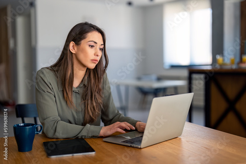 Serious millennial woman using laptop sitting at the table in a home office, typing, communicating online, writing emails, distantly working or studying on computer at home.